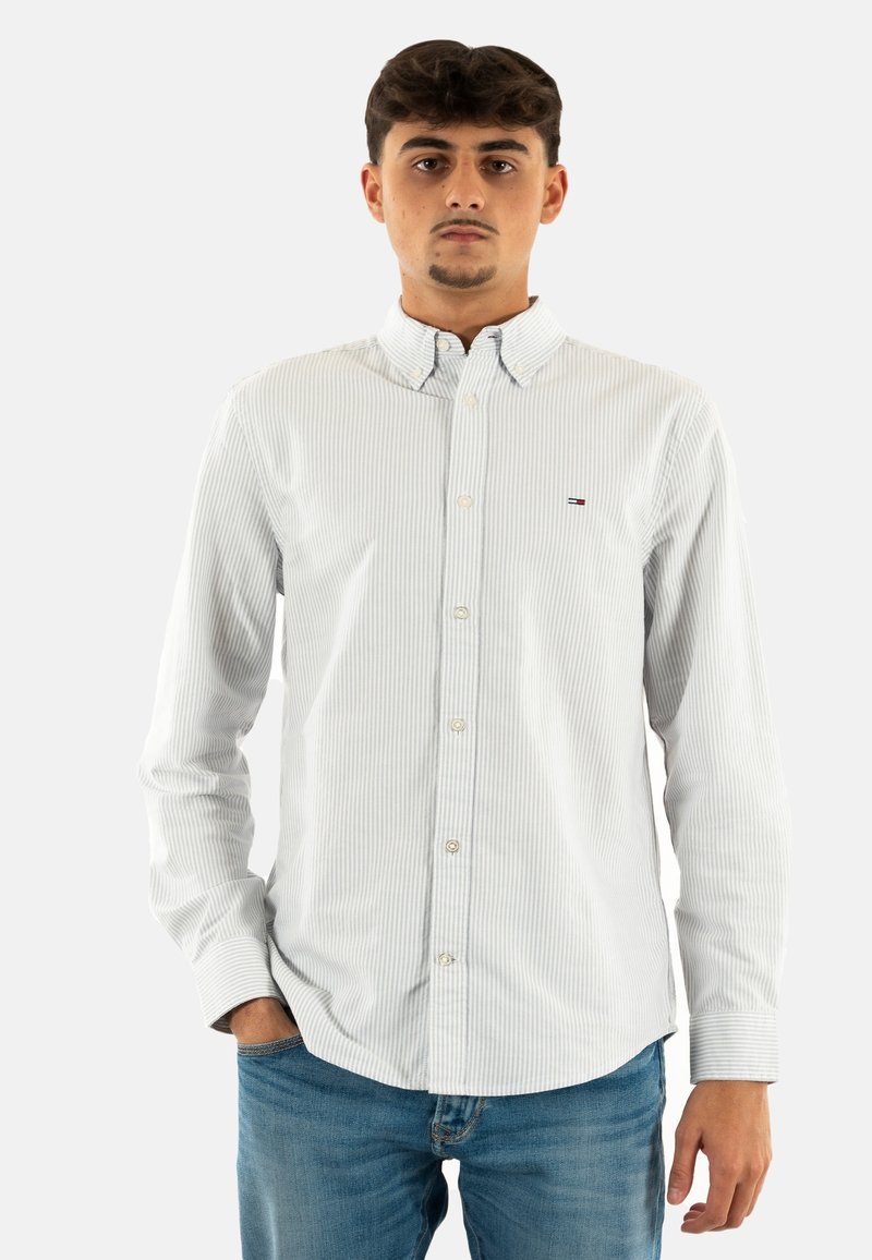 Camicia bianca a righe con colletto a punta. Presenta una placca con bottoni e un piccolo logo vicino al petto. Abbinata a jeans blu.