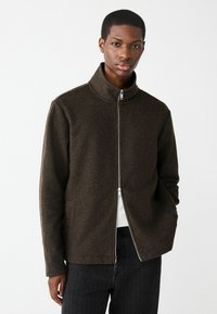 Hnedá zipsová bunda vyrobená z textúrovaného materiálu, s vysokým golierom a rovno strihaná. Nosená cez bielu košeľu a doplnená tmavými nohavicami.