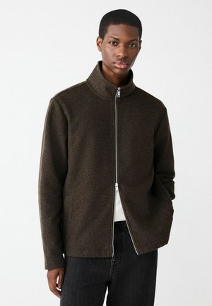 Braune Zip-Jacke aus einem strukturierten Stoff, mit hohem Kragen und geradem Schnitt. Getragen über einem weißen Hemd und kombiniert mit dunklen Hosen.