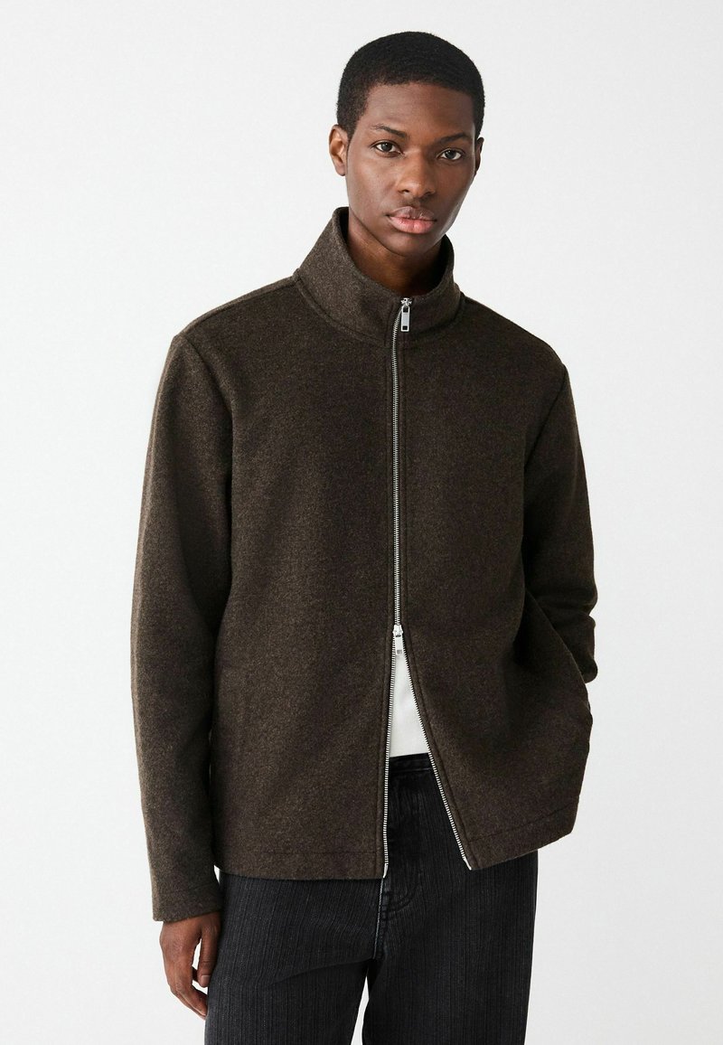 Hnedá zipsová bunda vyrobená z textúrovaného materiálu, s vysokým golierom a rovno strihaná. Nosená cez bielu košeľu a doplnená tmavými nohavicami.