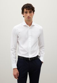Giovane uomo che indossa una camicia bianca aderente e pantaloni a righe blu scuro con una cintura nera, in piedi con una mano in tasca.