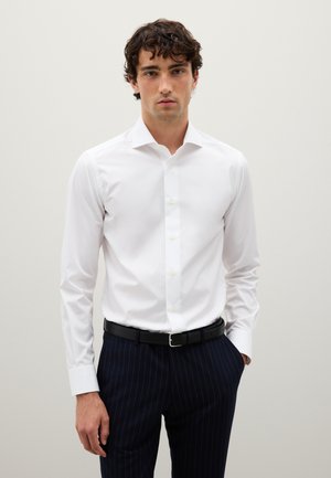 Junger Mann in einem figurbetonten weißen Hemd und dunkelblauen Nadelstreifen-Hosen mit einem schwarzen Gürtel, der mit einer Hand in der Tasche steht.
