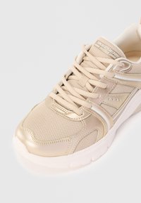 Beige ja metallikulta Skechers-sneakerit, joissa on verkkokangas, nauhakiinnitys, pehmustettu varsi ja paksu valkoinen pohja.