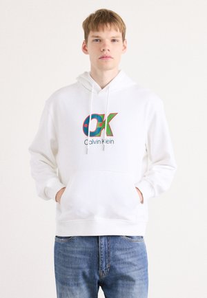 Biela bavlnená mikina s prednou vreckom. Obsahuje dekoratívne logo "OK" v pestrých farbách a text "Calvin Klein". Voľný strih s kapucňou na šnúrku. Modré džínsy.