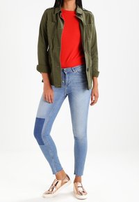 Olijfgroene jas met twee voorzakken, rode top, lichtblauwe skinny jeans met een donkere vlek en metallic roségouden instappers.