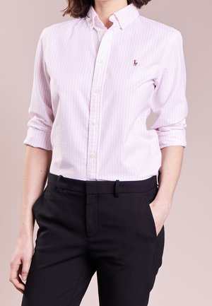 Kobieta w jasnoróżowej koszuli w paski z guzikami, rękawy podwinięte, czarne spodnie, jedną ręką w kieszeni, stojąca na neutralnym tle.