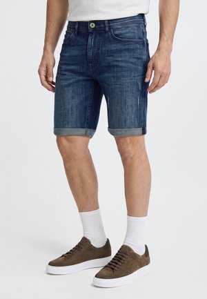 Un mannequin porte un short en jean bleu foncé à revers, une chemise blanche, des chaussettes blanches et des baskets marron sur un fond uni.