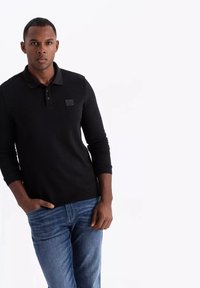 Svart långärmad pikétröja med en texturerad tyg och knäppning. Har en liten logotyp på bröstet, tillsammans med blåjeans.