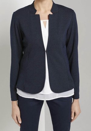 Femme portant un blazer bleu marine cintré avec un motif discret à pois sur un chemisier blanc avec une petite découpe triangulaire au niveau de l'encolure.