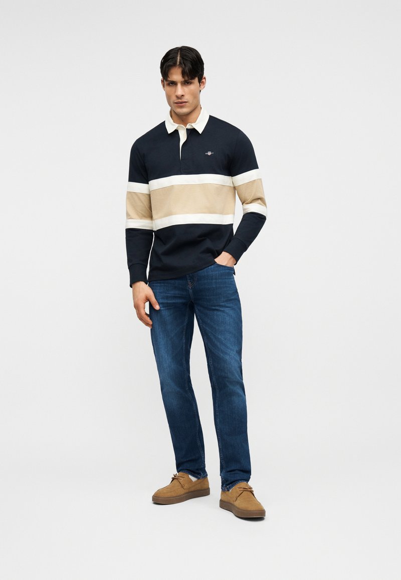 Homme adulte debout, portant un polo à manches longues rayé bleu marine, beige et blanc, un jean bleu et des chaussures en daim beige sur un fond clair uni.