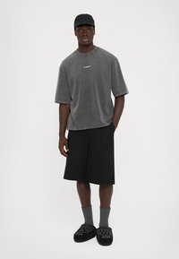 Man staat in een gewassen grijze oversized t-shirt, zwarte pet, zwarte knielange shorts, grijze sokken en zwarte bloemen slip-on schoenen.