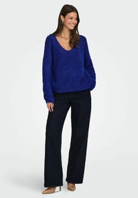 Prevelik modri pulover z teksturo in V-izrezom, v kombinaciji s temnimi ravnimi kavbojkami in nude visokimi petami. Ima dolge rokave in sproščen kroj.