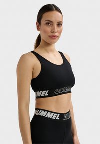 Vrouw met donker haar in een paardenstaart, die een zwarte sportbeha en leggings draagt met het "HUMMEL"-logo op elastische banden, tegen een effen achtergrond.
