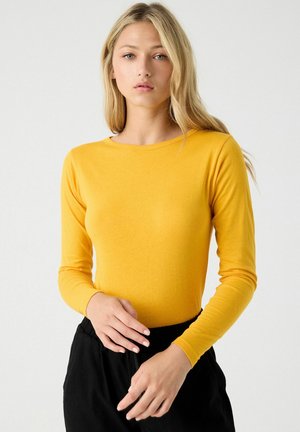 Top ajustado de manga larga en algodón amarillo brillante. Presenta un escote redondo y una textura suave, combinado con pantalones negros de cintura elástica.