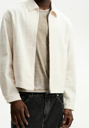 Mężczyzna ubrany w teksturowaną, jasnobeżową kurtkę zapinaną na zamek, pod którą ma beżowy T-shirt, oraz czarne jeansy, na jednolitym tle.