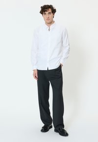 Mies seisoo päällään valkoinen nappipaita, mustat leveälahkeiset housut ja mustat loaferit valkoista taustaa vasten.