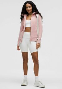 Svijetloružičasta zip-up dukserica s kapuljačom, uparen s bijelim sportskim grudnjakom i bijelim kratkim hlačama visokog struka. Teksturirani materijal; atletski dizajn.