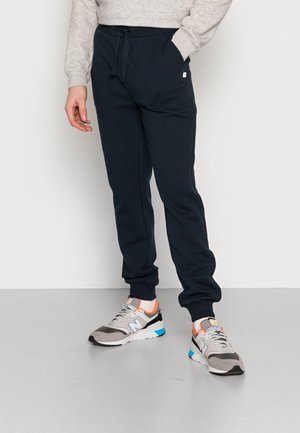 Person trägt marineblaue Jogginghose mit Kordelzugbund und weiße Sneakers mit orange- und blauen Akzenten, steht vor einem einfarbigen Hintergrund.