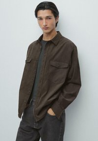 Młody mężczyzna o ciemnych włosach, ubrany w brązową koszulę zapinaną na guziki na ciemny t-shirt, czarne dżinsy, stoi z rękami w kieszeniach na jednolitym jasnym tle.