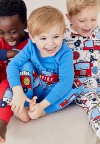 Trois enfants en pyjamas colorés : l'un en bleu avec des motifs de camions de pompiers, un autre en rouge, et un troisième en blanc avec des motifs de véhicules de police.