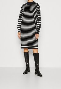 Raitapintainen neulemekko tummansinisestä ja kermanvärisestä langasta, pitkät hihat, päällysmallinen kaulus; yhdistetty mustiin nahkaisiiin polvisukkiin, joissa on korko.