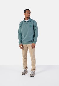 Sudadera de forro polar verde con cremallera y logo en el pecho, combinada con pantalones beige y zapatillas grises, que presenta un corte relajado y puños acanalados.
