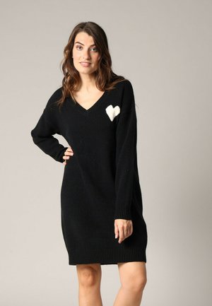 Robe en tricot noire avec un décolleté en V, des manches longues et des poignets côtelés. Présente un design de cœur blanc sur le côté gauche de la poitrine. Texture douce.
