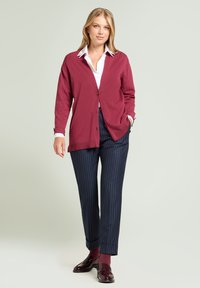 Burgundowa dzianinowa kardigan z przodem zapinanym na guziki, noszony na białej koszuli z kołnierzykiem. W połączeniu z granatowymi spodniami w prążki i ciemnymi mokasynami.