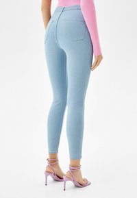 Jeans ajustados de mezclilla en un tono azul claro, con dos bolsillos traseros y una cintura alta. Combinados con sandalias de tacón alto en color lavanda.