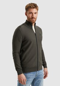 Temnozeleni zip-up pulover z rebrasto teksturo, visokim ovratnikom in logotipom na levi prsni strani. Nosi se nad svetlo srajco in v kombinaciji z modrimi kavbojkami.