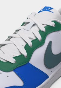 Baskets blanches Nike avec des accents en cuir vert et bleu, des lacets blancs, et un logo Nike bleu visible sur la languette.