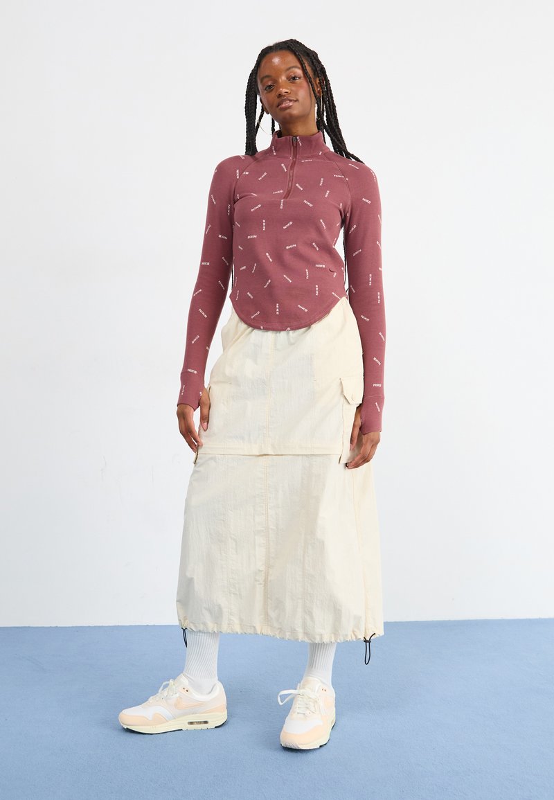 Haut à manches longues bordeaux avec un col haut et un motif de texte blanc, associé à une jupe portefeuille de couleur crème et des baskets claires.