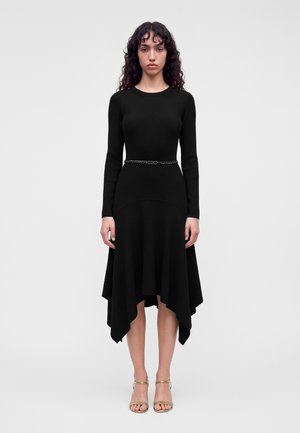 Femme aux cheveux bouclés et foncés portant une robe noire ajustée à manches longues, ourlet asymétrique, fine ceinture noire et talons ouverts dorés.