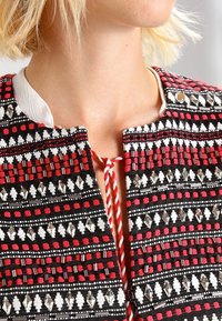 La veste brodée noire, blanche et rouge présente un motif texturé avec des perles décoratives et une fermeture à cordon tressé au niveau du col.