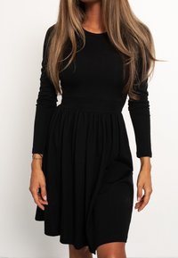 Robe noire à manches longues avec un corsage cintré et une jupe évasée. Confectionnée dans un tissu doux, elle présente des fronces au niveau de la taille.