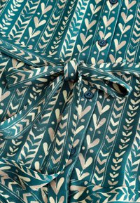 Lätt, mönstrad tygskjorta i teal, med krämfärgade blad- och hjärttryck, knytning i midjan och mörka knappar.