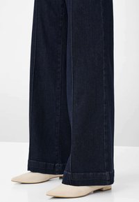 Jeans larghi a gamba larga di colore blu scuro abbinati a scarpe beige a punta con tacco basso su un pavimento bianco con sfondo semplice.