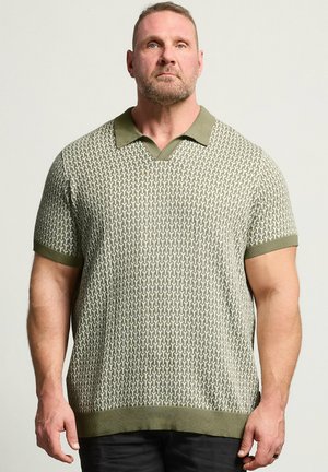 Muskuløs mand med kort hår og skæg iført en grøn og beige mønstret kortærmet poloshirt mod en ensfarvet baggrund.