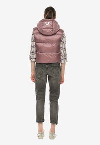 Vycpaná růžová vesta s kapucí, která má lesklou texturu. Modelka má pod ní na sobě kostkovanou košili a šedé džíny s ohrnutými nohavicemi.