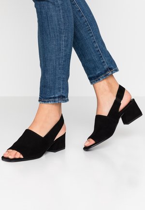 Pieds portant des sandales noires à talon bloc ouvert à l'avant avec brides à la cheville, associées à un jean bleu sur fond blanc uni.