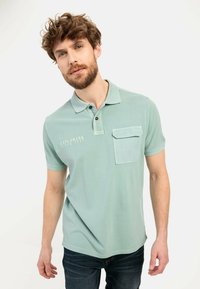 Lichtgroene polo met een gestructureerde zak, drie knopen aan de hals en een bedrukt logo. Draagt bij donkere jeans tegen een neutrale achtergrond.