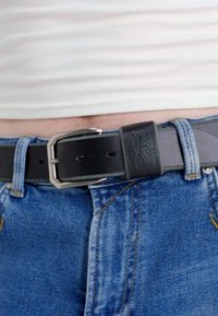 Cintura in pelle nera con fibbia in metallo argentato, caratterizzata da una fascia testurizzata e cuciture visibili, indossata con jeans blu.