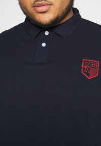 Navy polo shirt made of cotton, featuring a three-button collar and a red embroidered badge with geometric patterns on the chest.