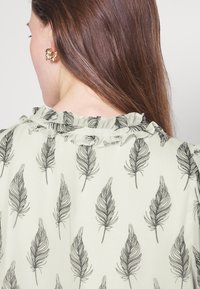 Blouse vert clair avec imprimé de plumes noires, dotée d'un col à volants et d'une texture lisse, partiellement montrée de dos.