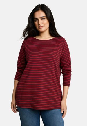 Frau mit langen dunklen Haaren, die ein rot-bordeaux gestreiftes Langarmshirt und blaue Jeans trägt, steht vor einem einfarbigen Hintergrund.