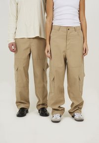 Khaki cargobroek met meerdere zijzakken, een relaxte pasvorm en een soepele stofafwerking. Draagt met zwart en grijs schoeisel.