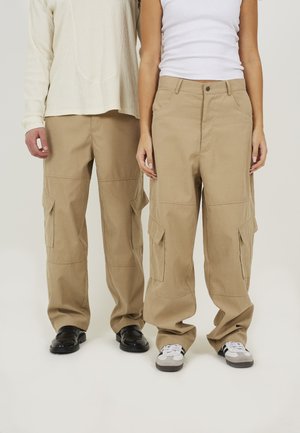 Khaki cargobroek met meerdere zijzakken, een relaxte pasvorm en een soepele stofafwerking. Draagt met zwart en grijs schoeisel.