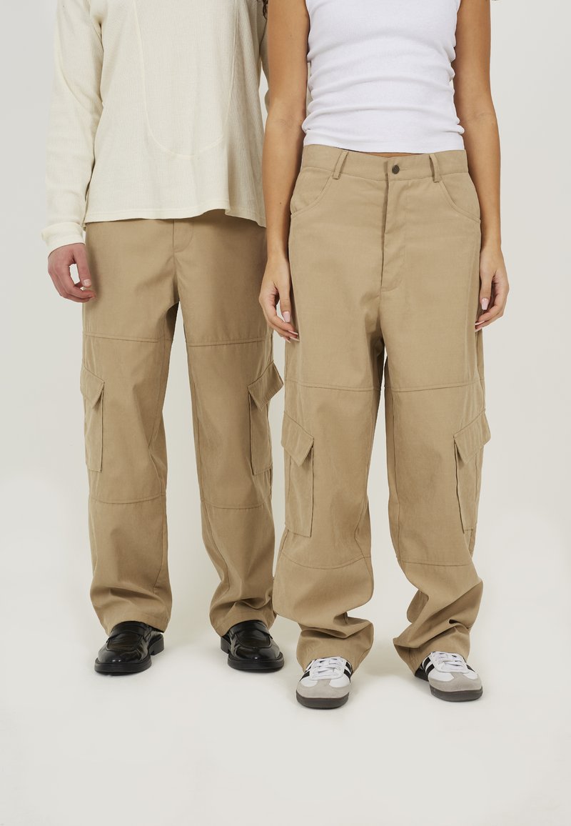 Khaki cargobroek met meerdere zijzakken, een relaxte pasvorm en een soepele stofafwerking. Draagt met zwart en grijs schoeisel.