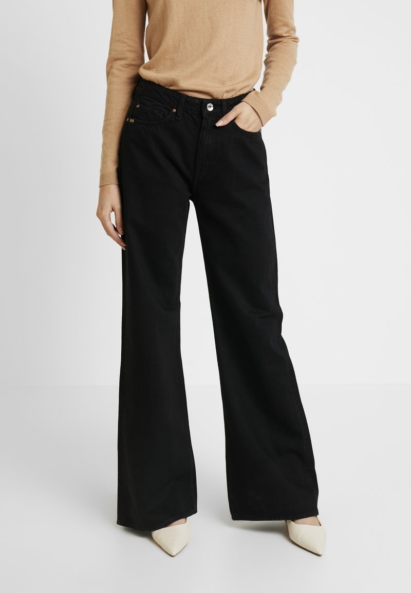 Henkilö, joka käyttää korkeavyötäröisiä leveälahkeisia mustia farkkuja, beigeä pitkähihasta toppia ja teräväkärkisiä valkoisia kenkiä, seisoo yksi käsi taskussa.