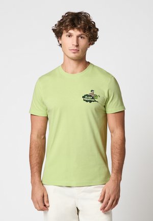 Jeune homme aux cheveux bouclés portant un T-shirt vert clair avec le motif "Plant Together" et un pantalon blanc, debout devant un fond clair uni.